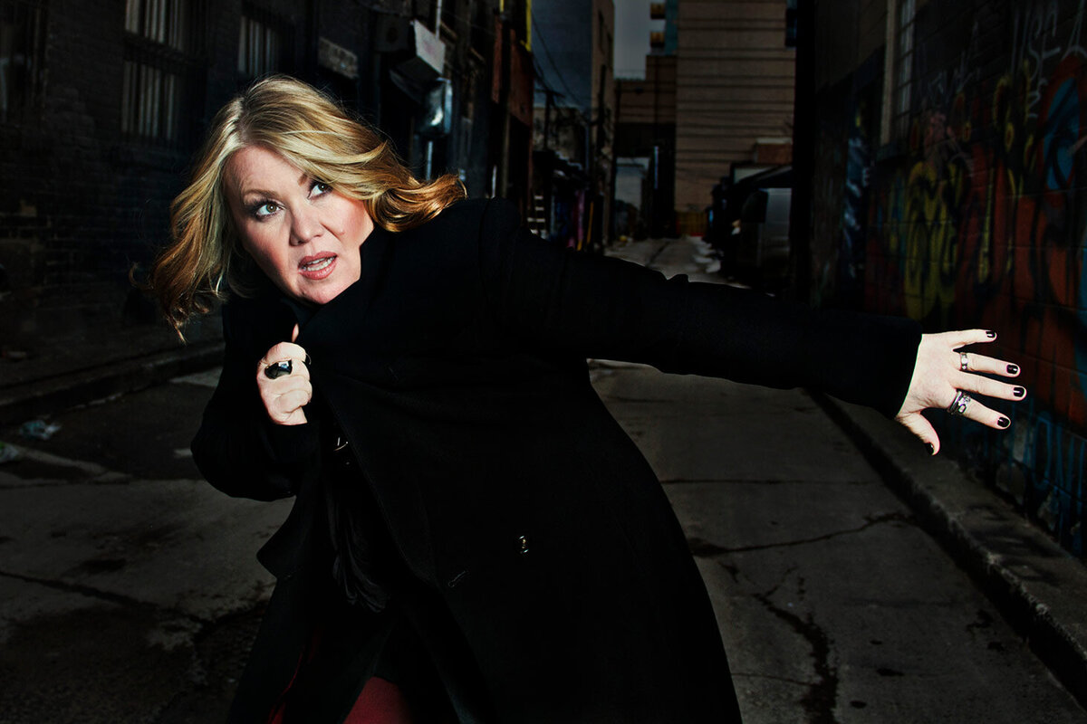 Jann Arden musician portrait clasping collar of black coat other arm outstretched while standing in alley