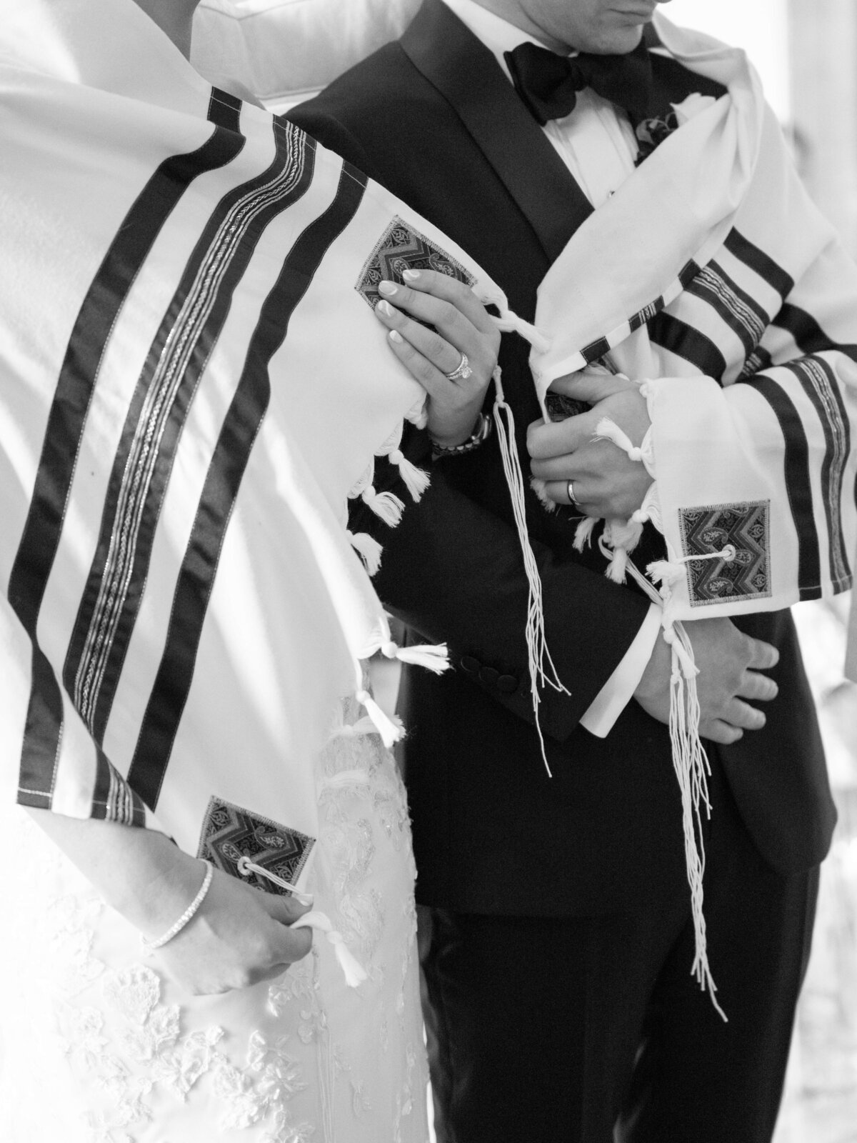 A black and white image of two people dressed in formal attire, wrapped in traditional Jewish prayer shawls with fringes and embroidered details during a ceremony. The focus is on the shawls and the hands holding them.