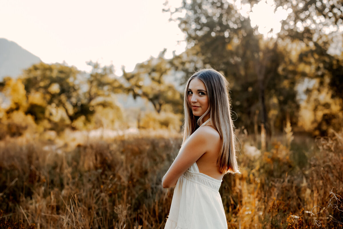 High school senior girl with long blond hair standing in a tall grassy field with rays of sunshine behind her