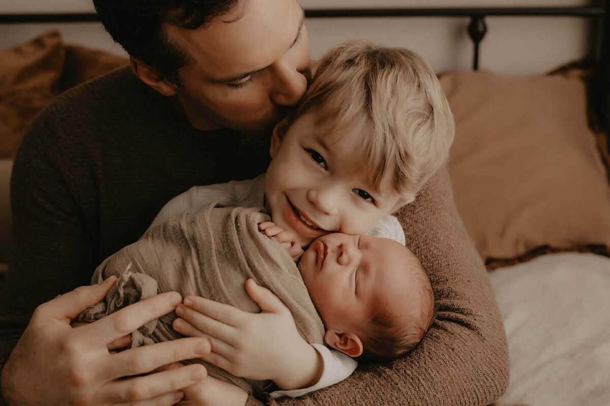 A father helps his son hold his new baby brother while the dad kisses the older boys head
