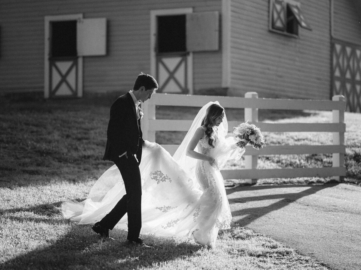 A groom holding a bride's train as they walk outside.