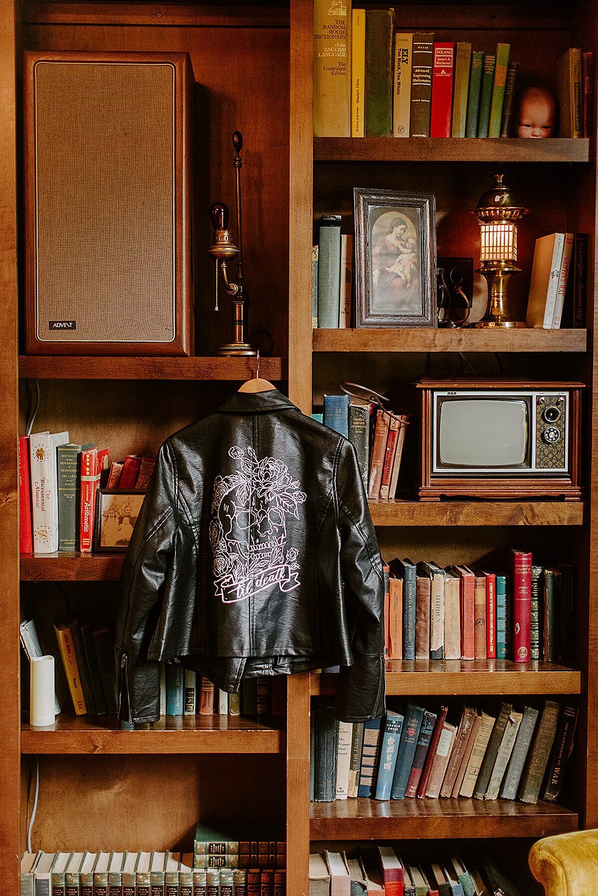 A large wooden bookcase filled with vintage books, a small TV and a speaker at Rosemary and Beauty Queen in Nashville has a bride's black leather jacket hanging from a wood handle. The back of the jacket is detailed with a floral skull drawn in white with "Till Death" in calligraphy at the bottom.