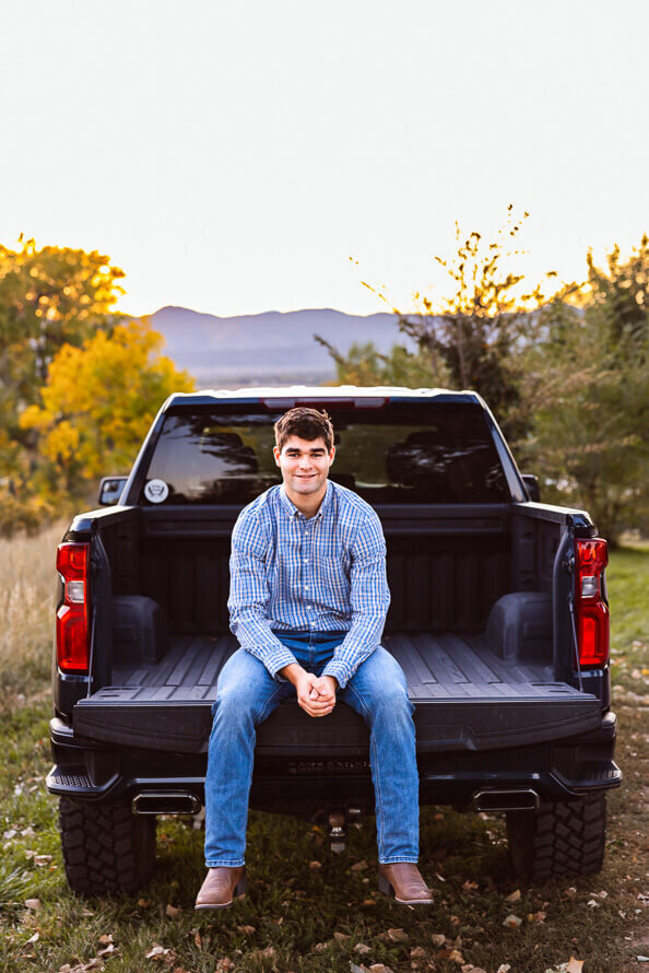 Boy sits on the back of his truck with mountains in the background.