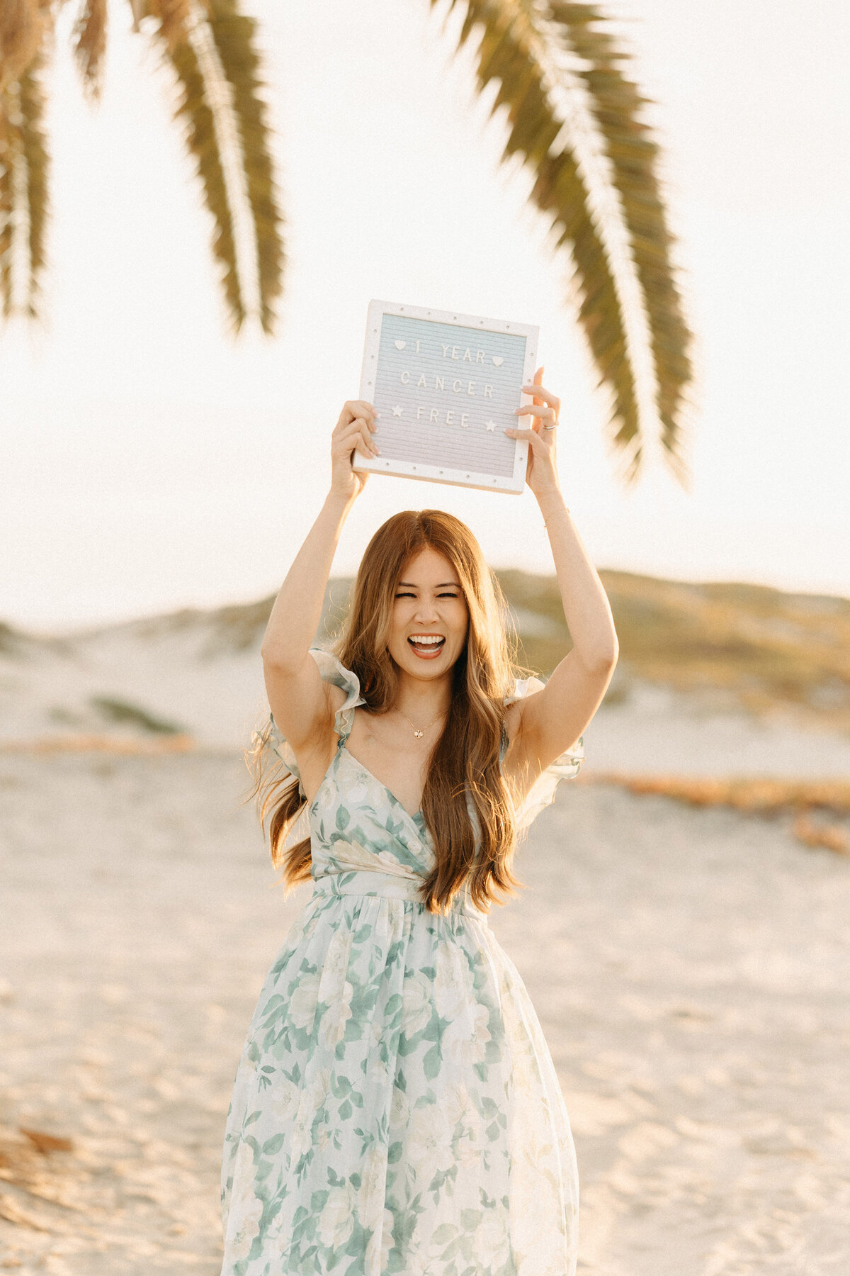 Smiling woman in a floral dress holding a sign above her head reading ‘1 year cancer free’ during a golden hour photoshoot on Coronado Beach
