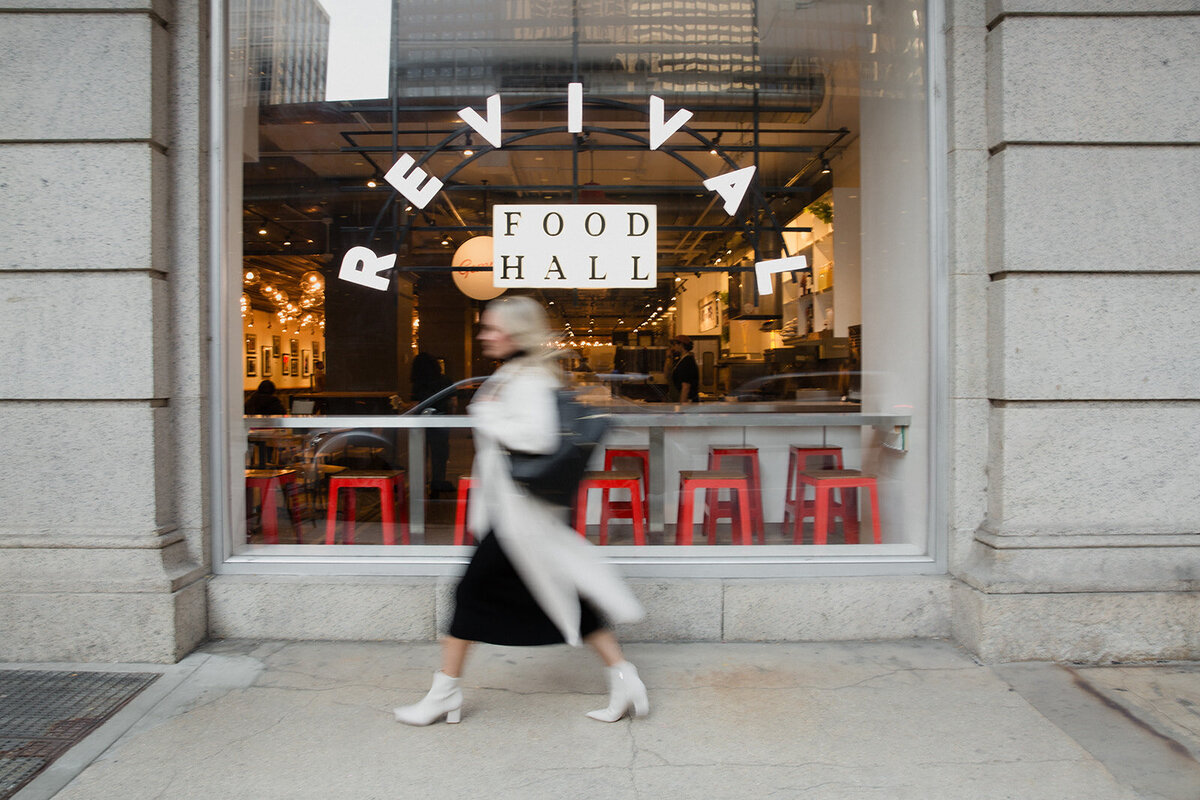 blonde woman walking past a restaurant in Chicago, she is blurred because she walks so fast. She wears a black dress, white boots and long white coat