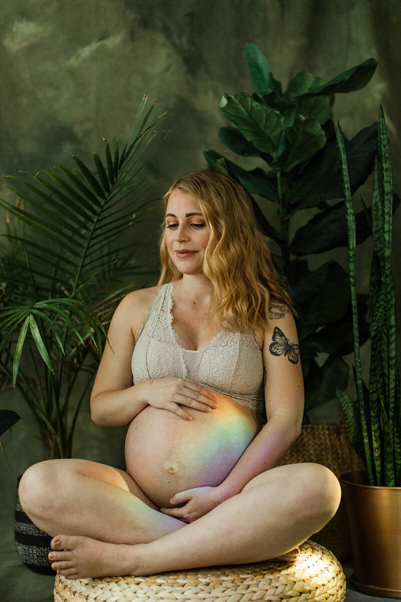 pregnant mom sits surrounded by house plants and rainbow projected on her stomach