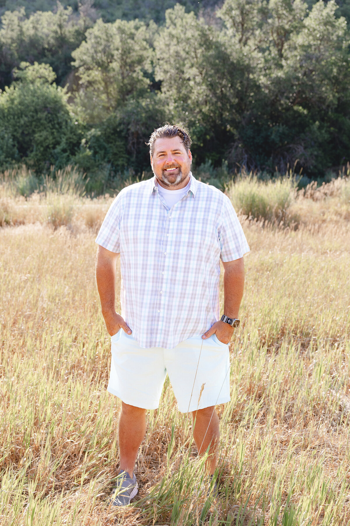A father with short, wavy hair and a beard smiles warmly, standing relaxed in a field with lush greenery behind him in Midway, Utah.