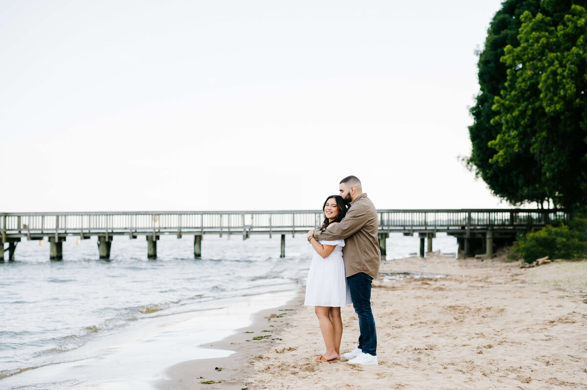 Virginia weddin gphotographer captures engagement photos on a beach at woodbridge lake with man standing behind woman and holding her around her shoulder and kissing her head with lush trees far from the water and a dock in the distance