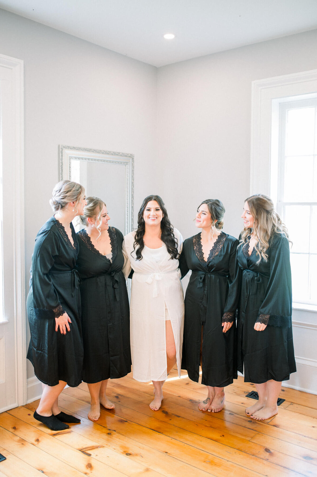Bride wearing white robe in the center of her bridesmaids wearing black robes