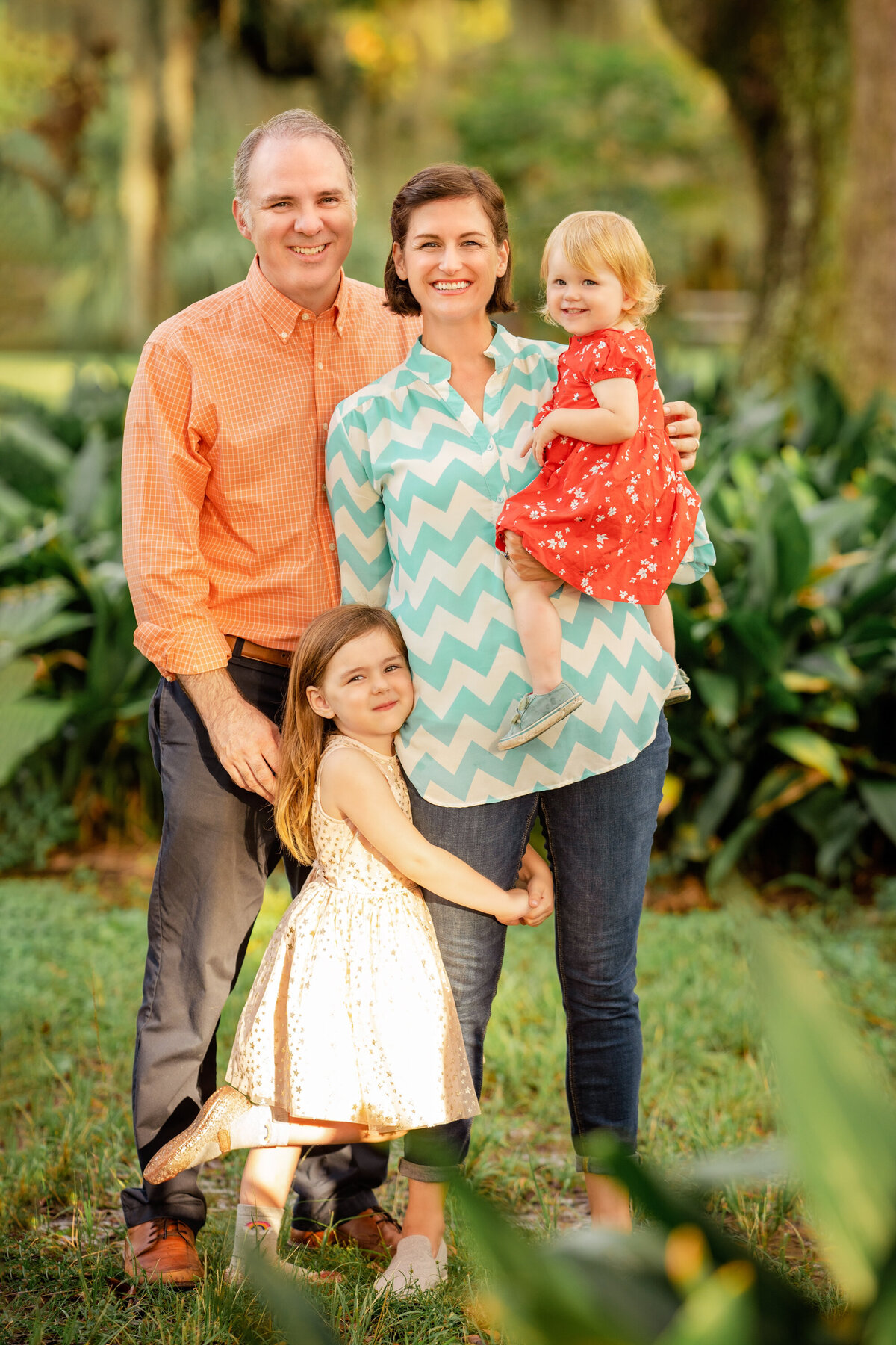 Family standing near the cast iron plants in City Park.  Dad is older, wearing an orange shirt.  Mom has short brown hair and is wearing an aqua and white chevron shirt.  The youngest daughter is wearing a red dress and is being held by mom.  The older daughter has her arms wrapped around mom's leg.