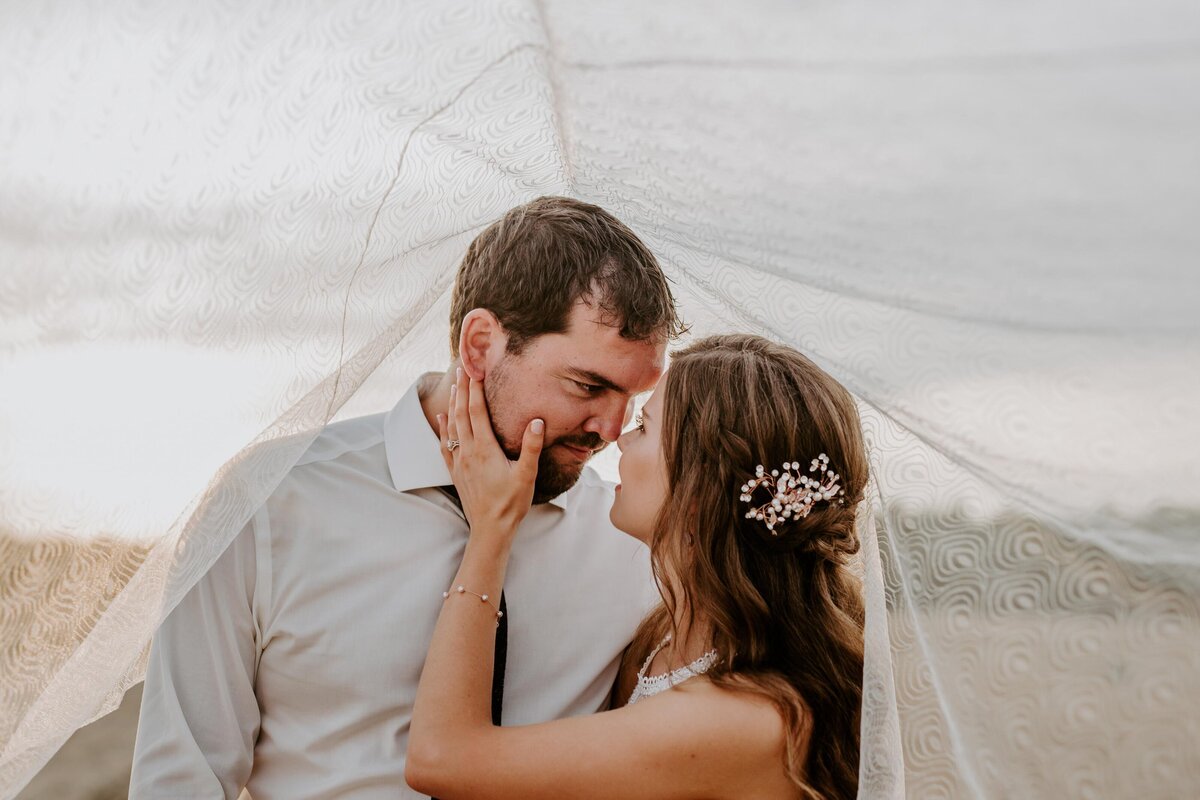 Bride and groom touching noses for wedding portrait. Bride's hand is resting on groom's cheek and her veil is blowing overtop of them.