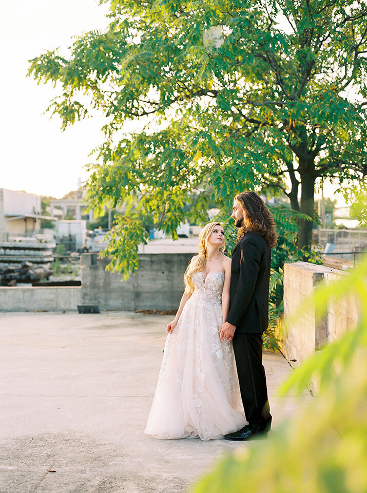 Bride and groom wearing a black tuxedo and white wedding gown looking at each other outside on a rooftop.
