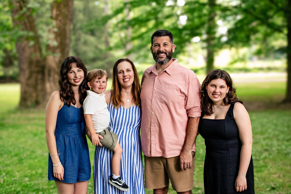 A family of five with red hair and dark features pose in a park with the green woods in the backdrop near Sewickley Pennsylvania