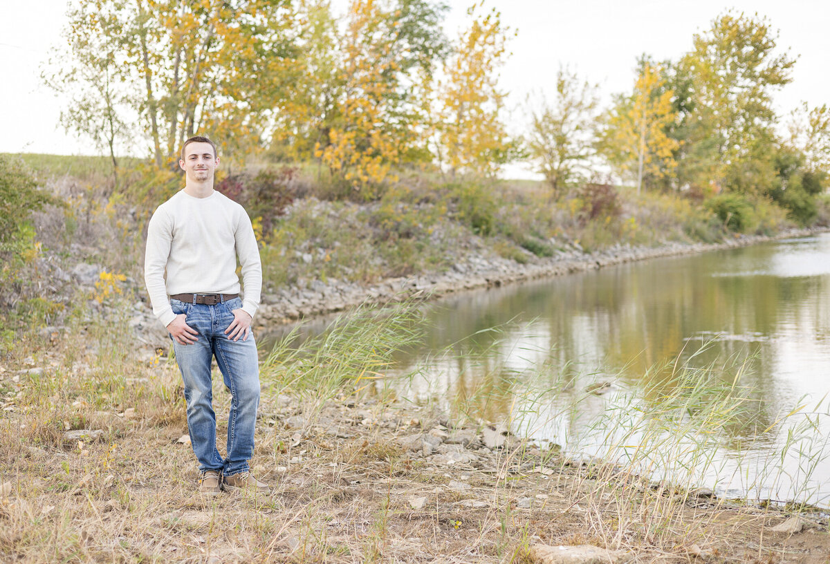 high school senior male, standing next to a pond, with trees in the background, thumbs in his pockets, wearing a light tan long sleeve shirt, blue jeans, a belt, and tan dress shoes, looking at the camera smiling
