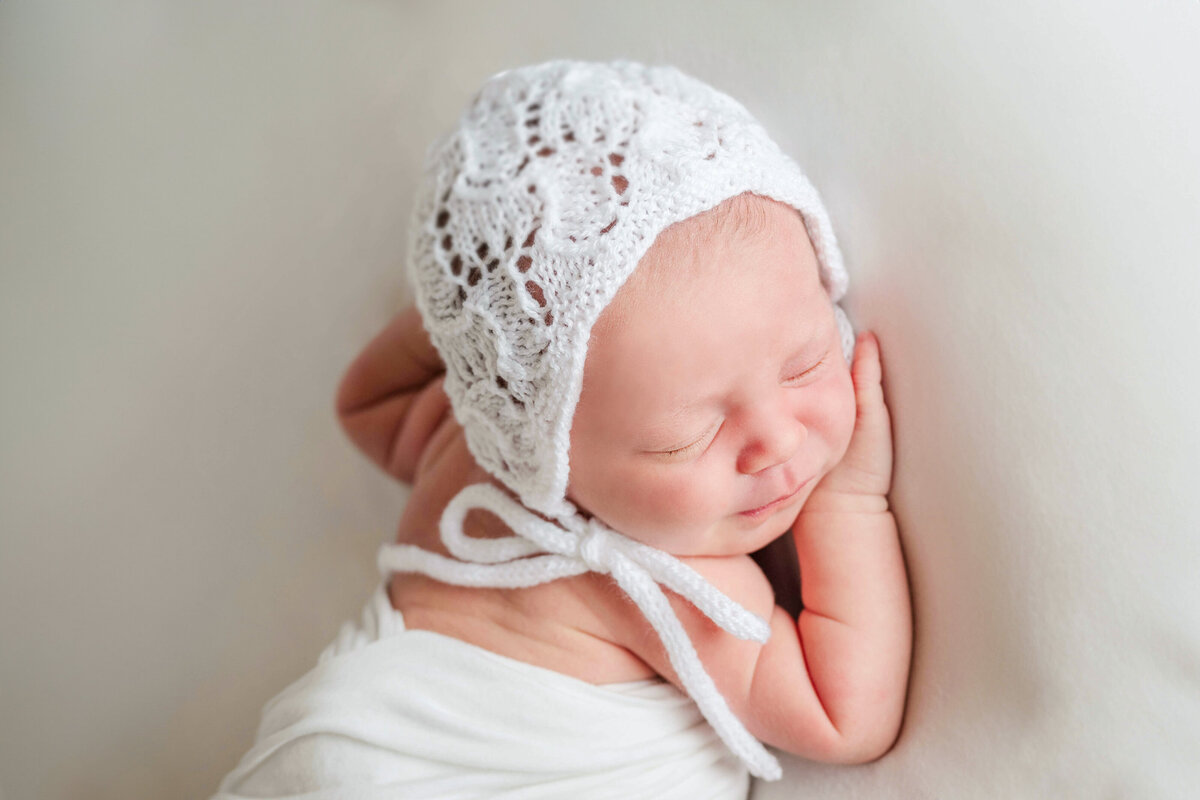 A newborn baby girl wrapped in a whie blanket and sleeping, while wearing a white knitted bonnet.