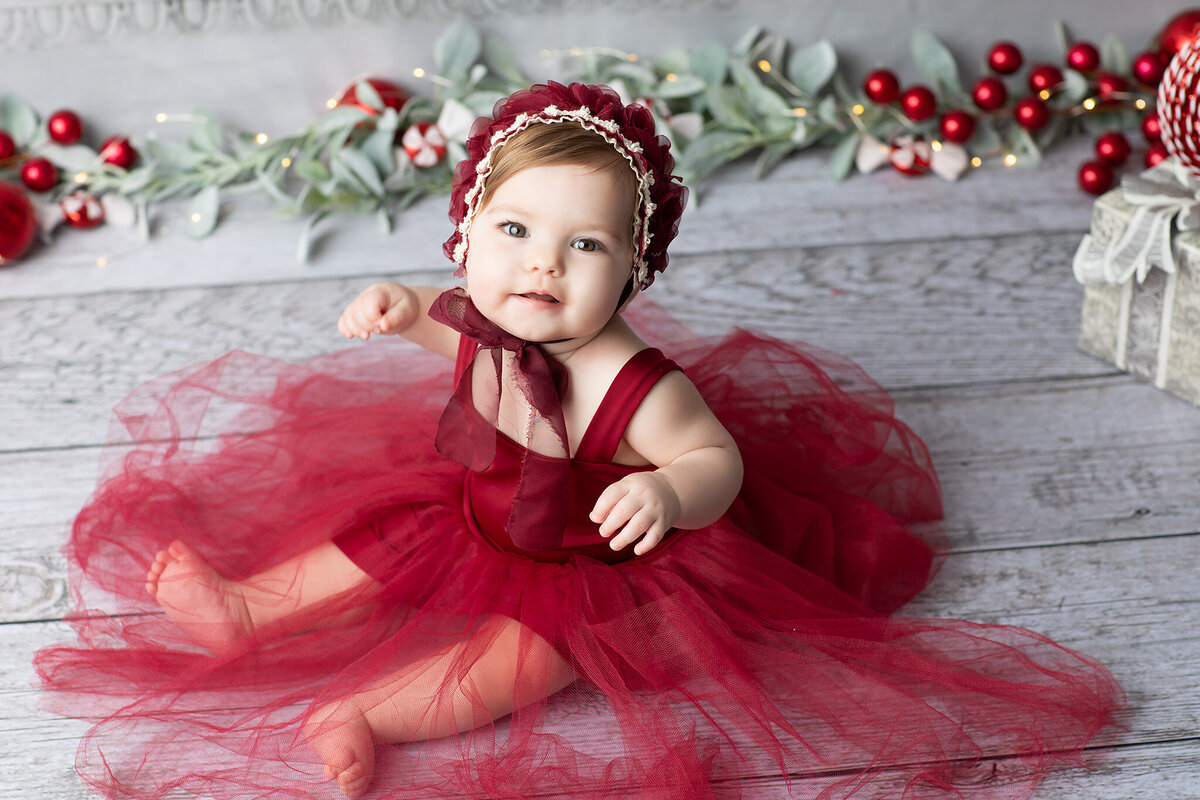 Smiling baby girl in a red holiday dress and matching bonnet poses on a wooden floor surrounded by festive garland and presents, captured by a Dallas milestone photographer.