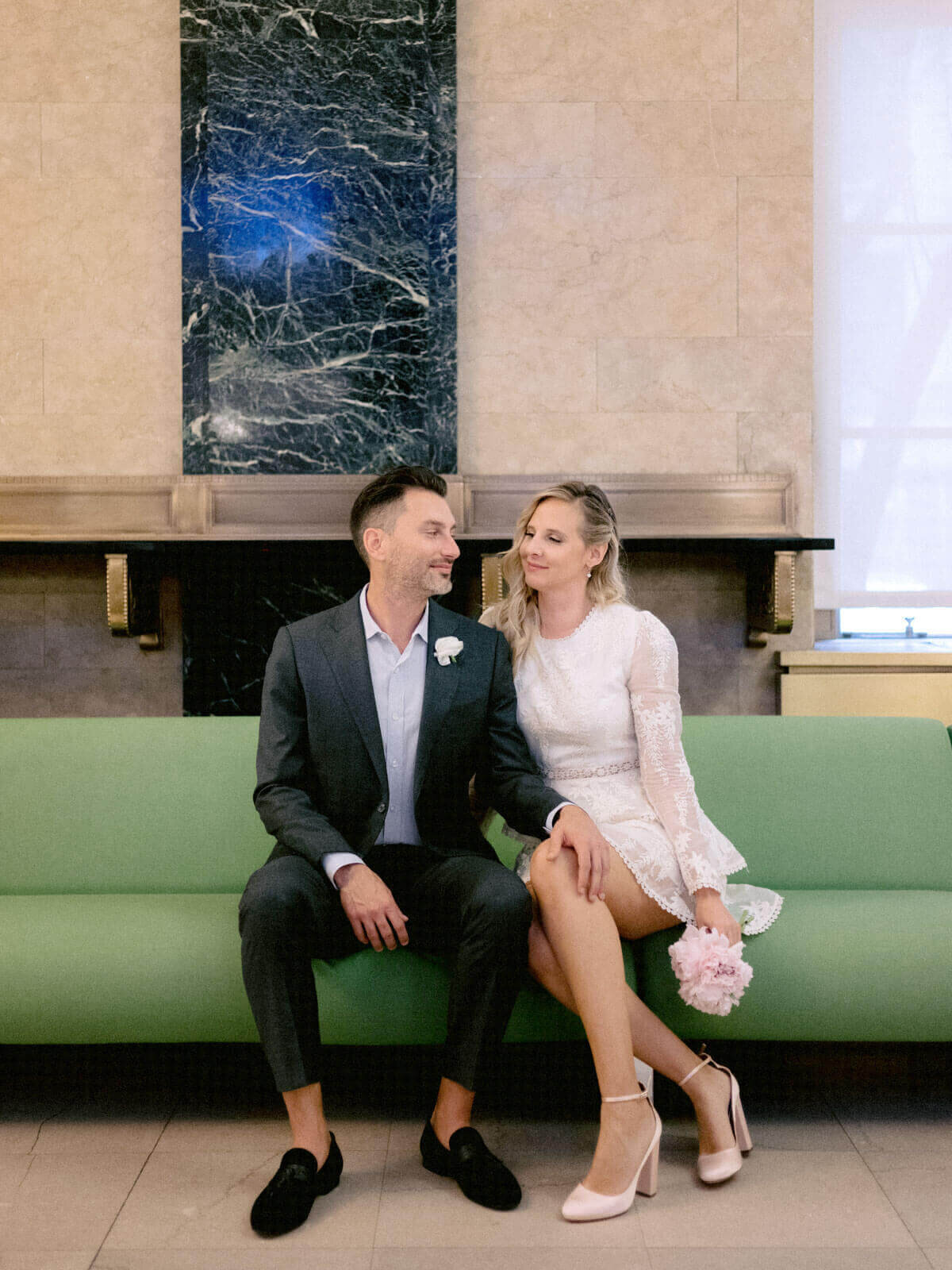 The bride and groom are waiting for their turn on a green couch in NY City Hall for their elopement. Image by Jenny Fu Studio