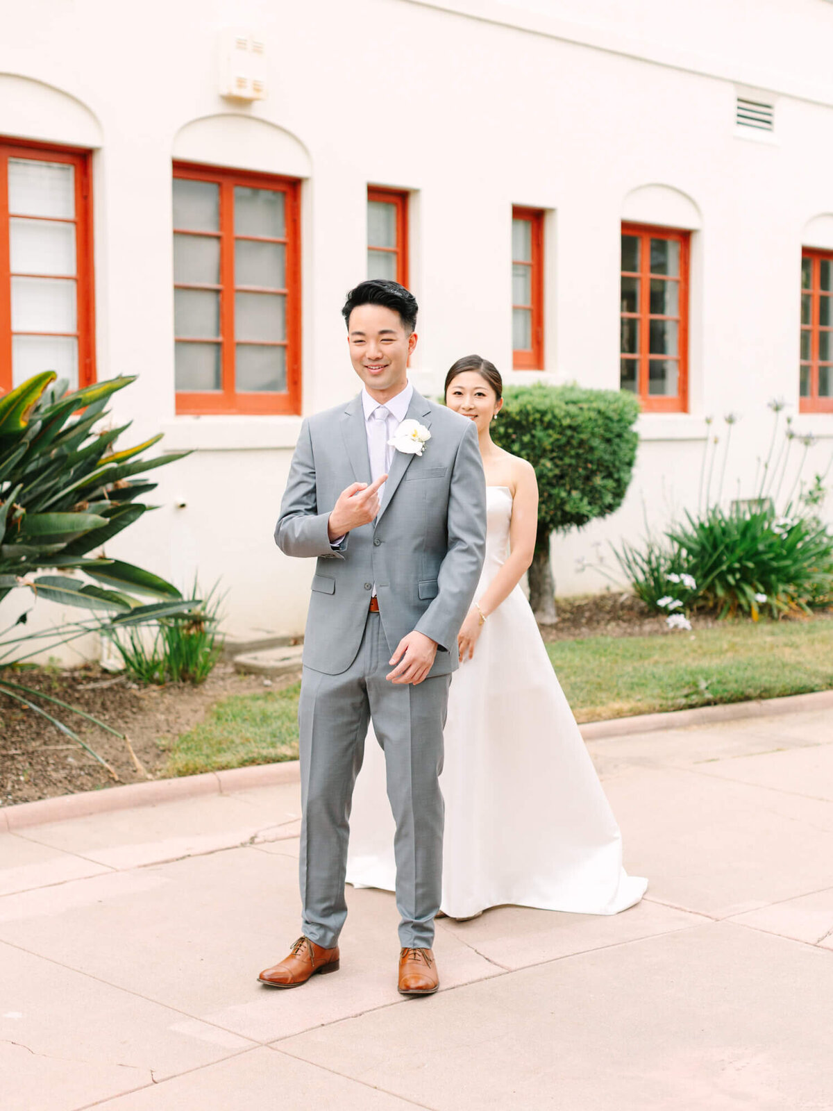 A groom in a gray suit smiles as a bride in a white gown stands behind him, outside a building with red-framed windows and green shrubs, exuding joy.