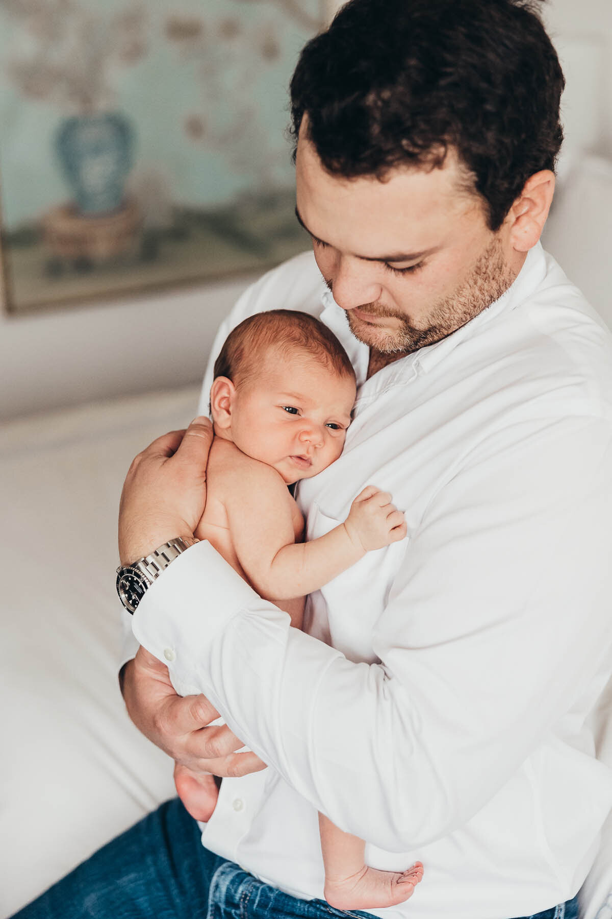 a father in a white shirt holds his newborn baby girl against his chest in their san diego home