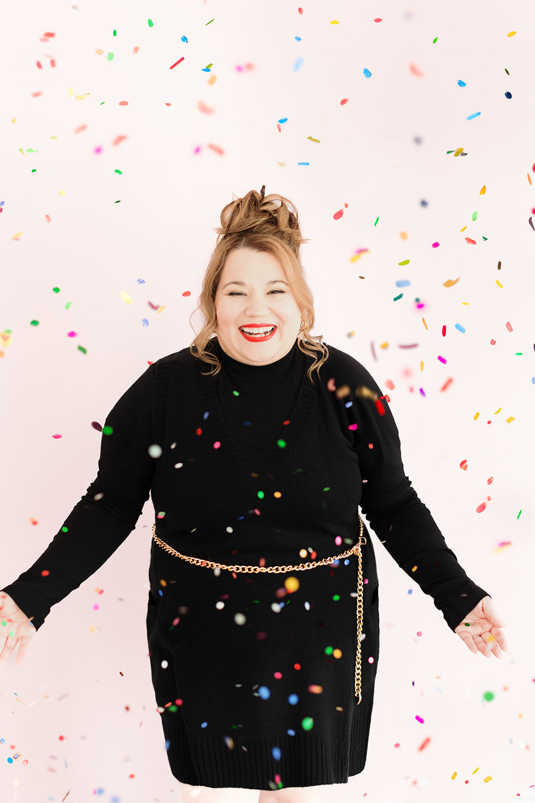 joyful woman in black dress in front of pink backdrop standing in a flurry of confetti and laughing