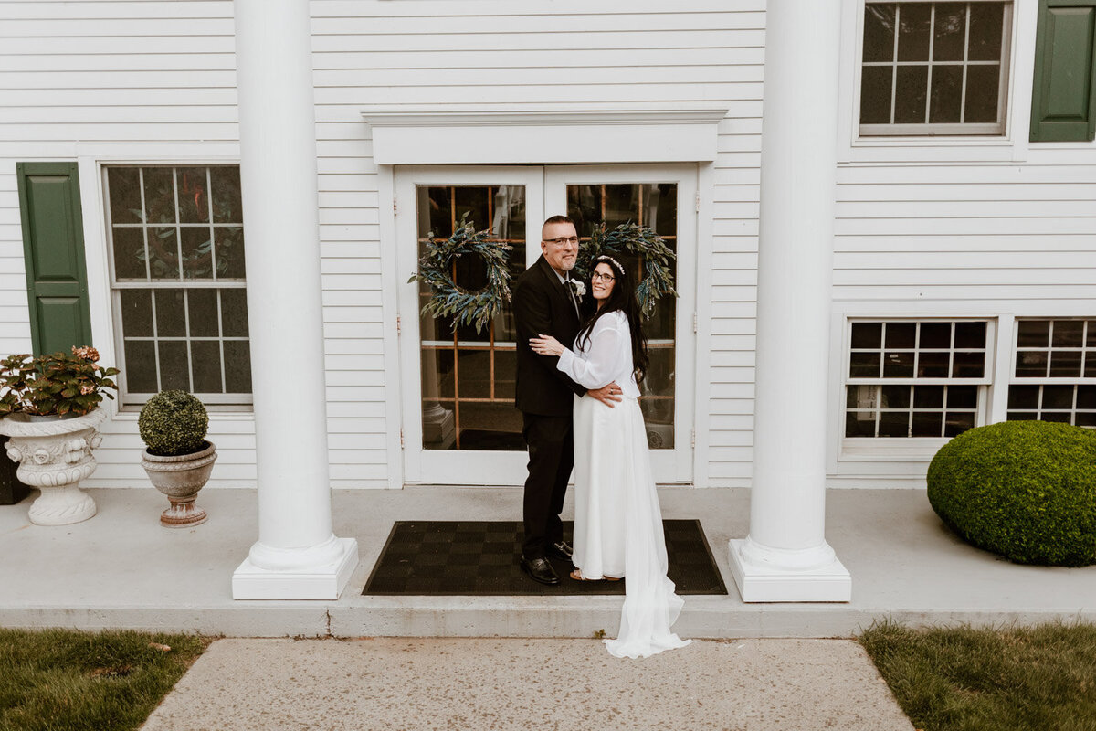 Hudson Valley wedding photographer captures a loving couple's embrace at the Curry Estate, framed by the venue's iconic white colonial facade and festive green wreaths.