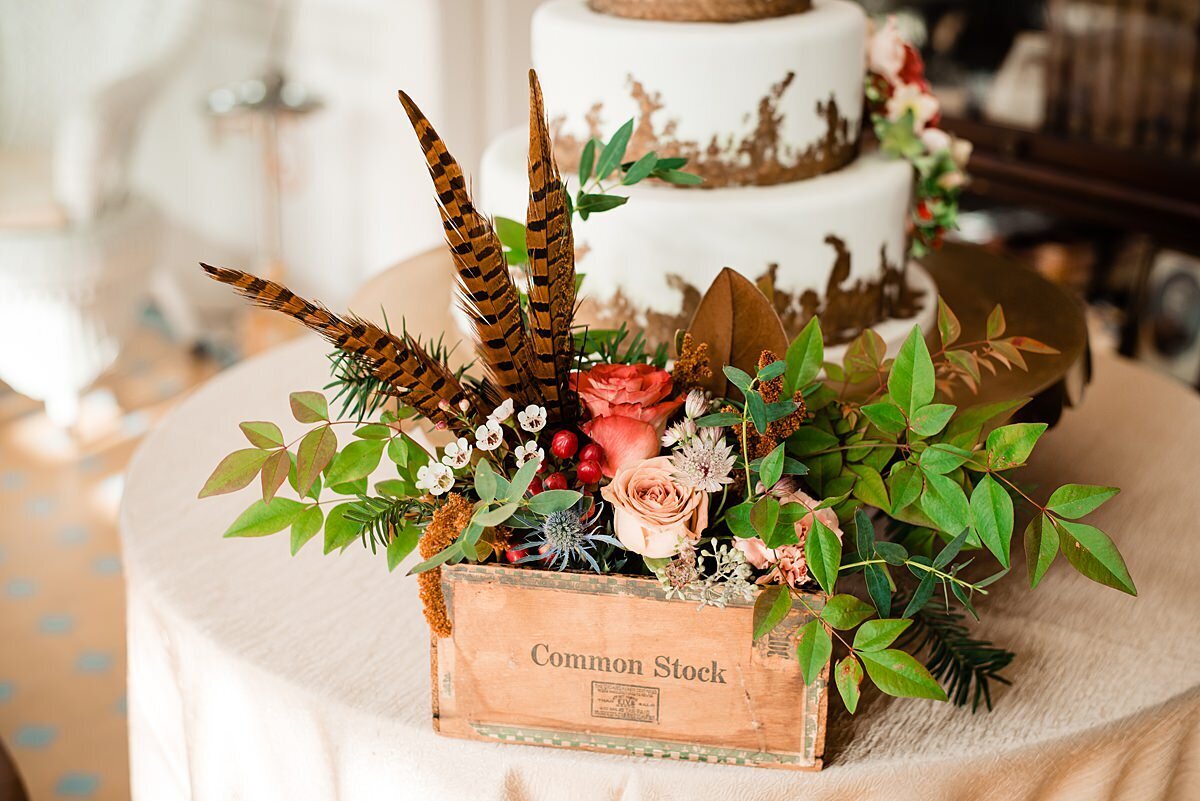 A square wooden cigar box sits on a table covered with a light gold table cloth. The cigar box has brown and black feathers, magnolia leaves, assorted greenery with orange roses, blush roses and chamomile in a centerpiece in front of a three tier white wedding cake with gold leaf at Rippa Villa.