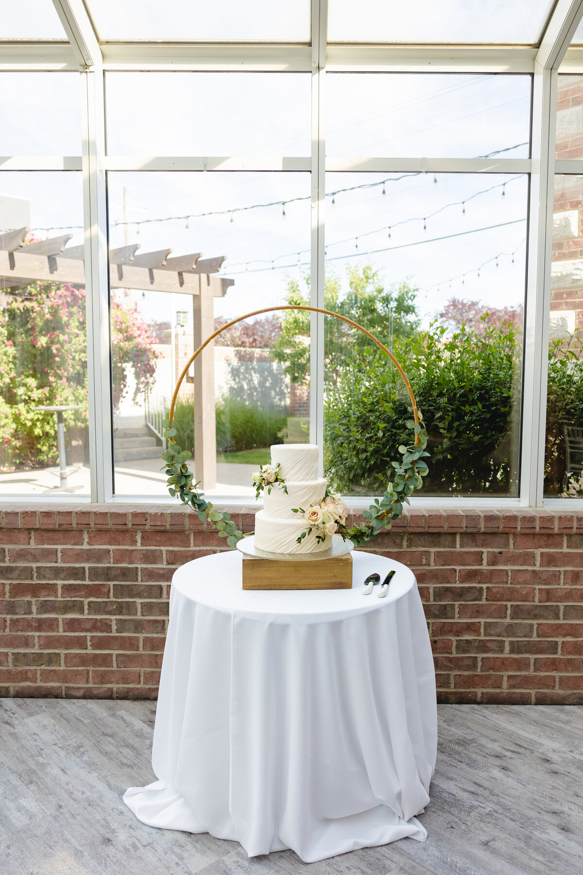A three-tiered white wedding cake with soft floral decorations, displayed under a circular greenery arch inside a sunlit reception space.