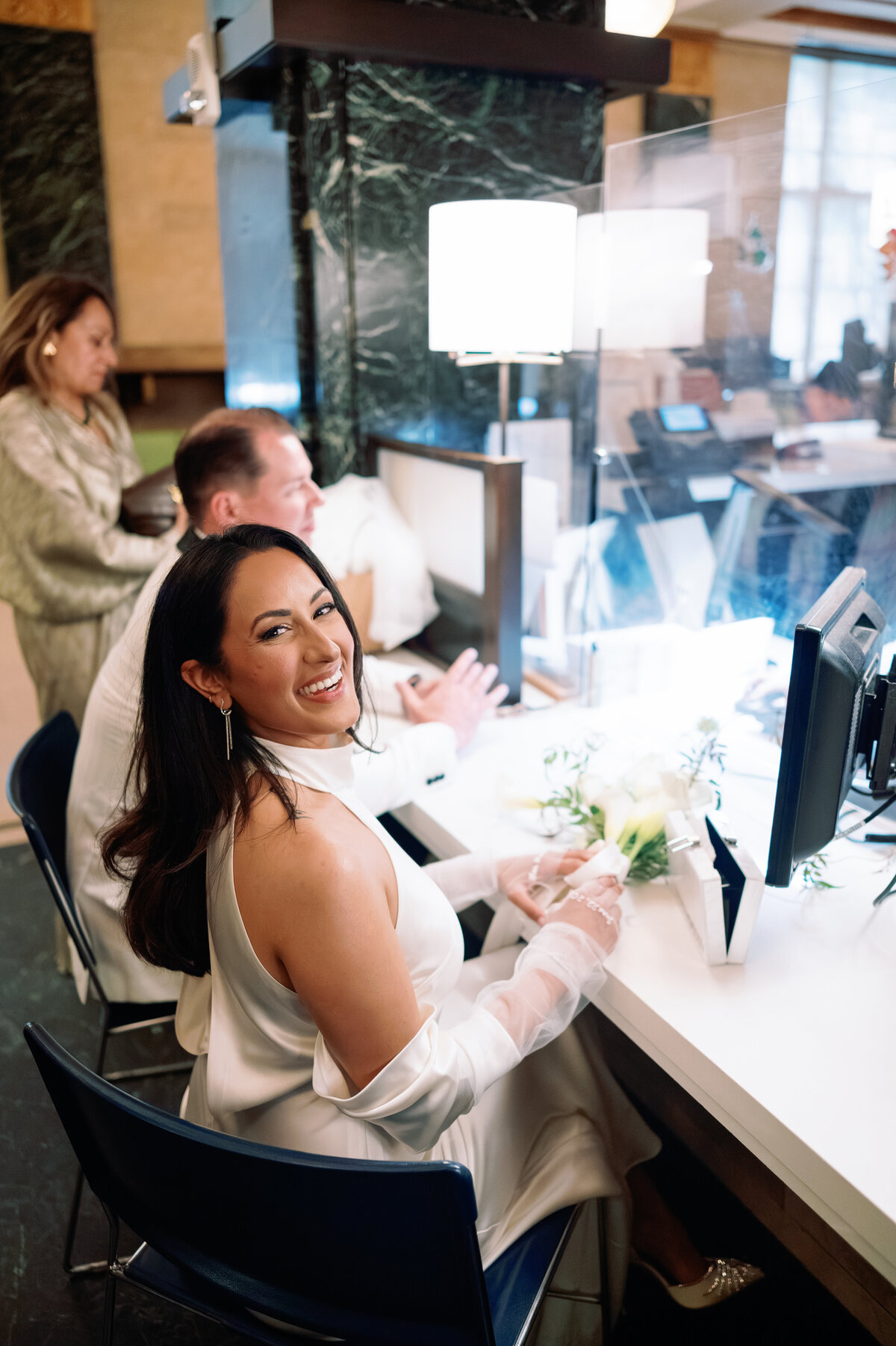 Bride smiling while sitting at the marriage license desk inside New York City Hall during her elopement with Chris, captured candidly by NYC wedding photographer Perry Hancock.