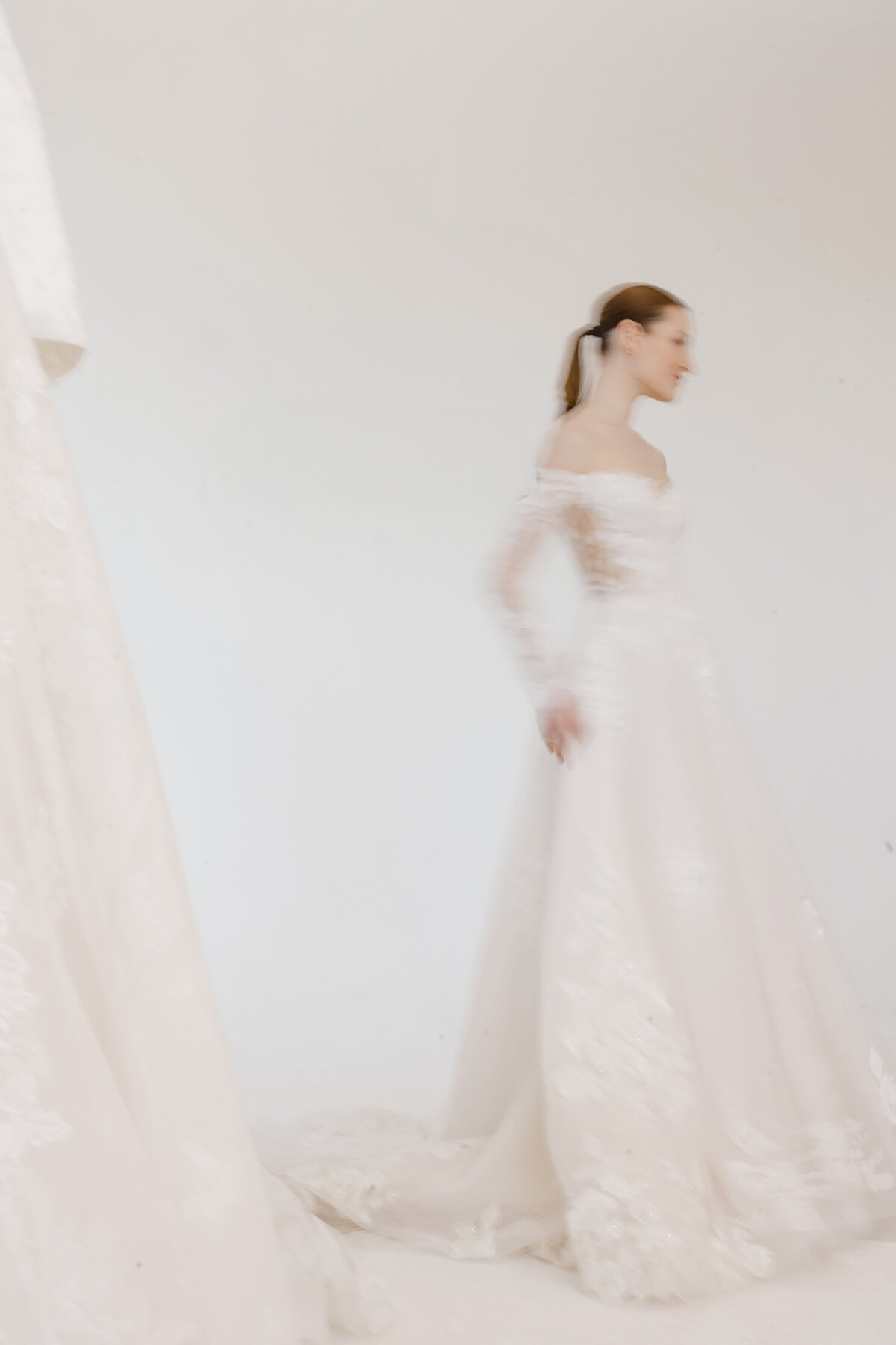Motion-blurred portrait of a model in a lace off-the-shoulder wedding gown walking across a clean white backdrop, photographed at the Museum of Natural History to highlight fluid movement and couture fabric details in a modern editorial style.