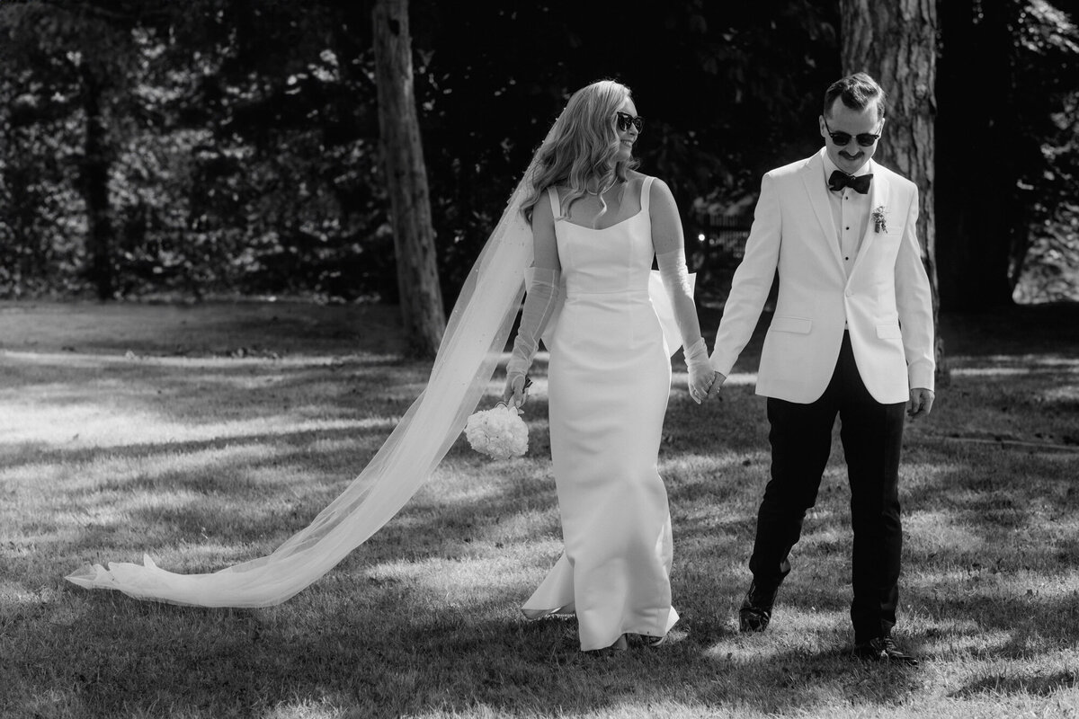 Black and white image of the wedding couple with sunglasses walking across the grass, the bride's long veil flies to the side.