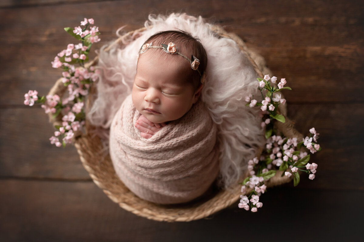 Baby girl wrapped in a blush knit swaddle for Jupiter and West Palm Beach newborn photoshoot.  Baby is sleeping in a woven basket atop a long furry rug. Baby's hands are peeking out of the swaddle and is wearing a delicate floral headband.