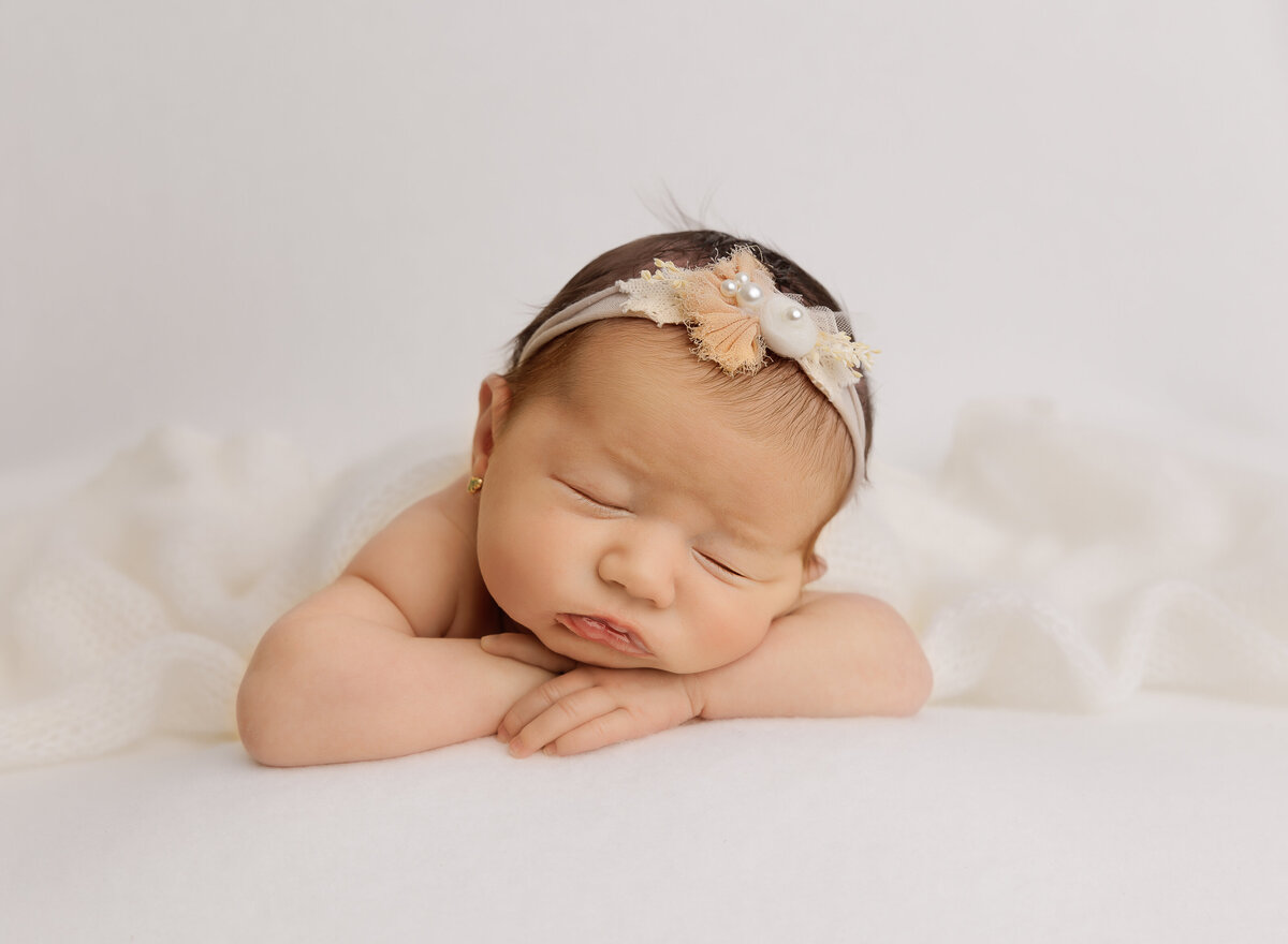 Baby girl is sleeping at her brooklyn newborn photoshoot. Baby's hands are folded under her chin and she is wearing a delicate floral headband. Captured by top Brooklyn newborn photographer Chaya Bornstein