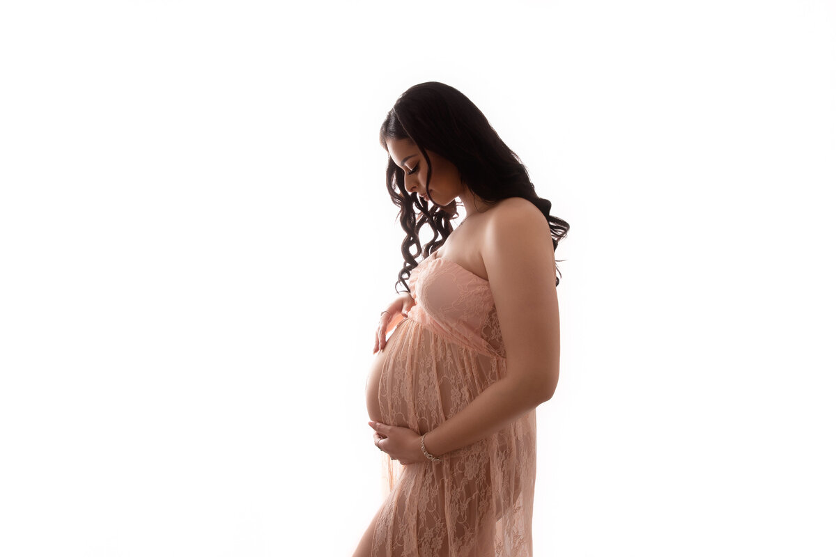 Pregnant woman wearing a peach lace dress and lovingly looking down at her belly.