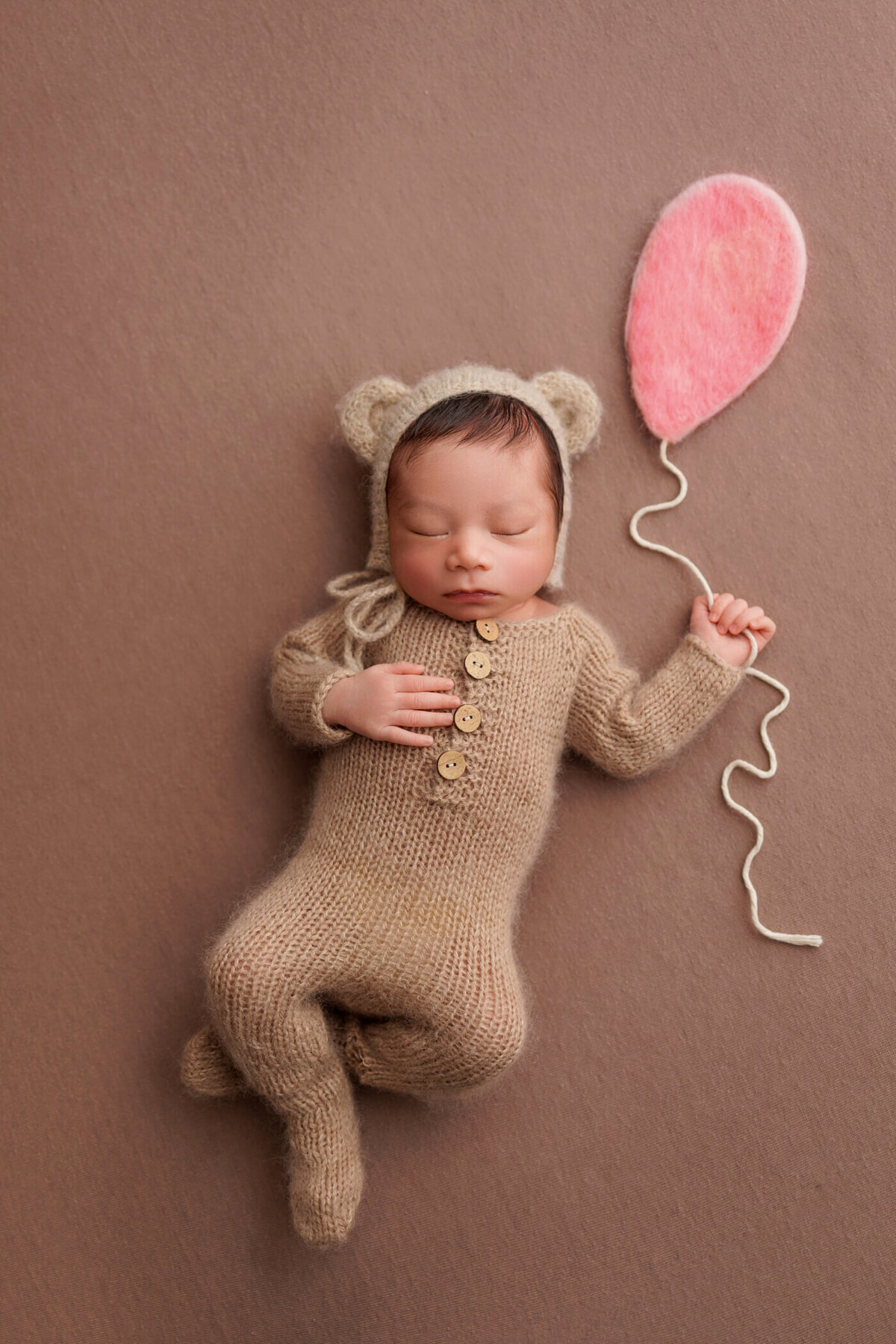 Newborn baby dressed in a knitted bear costume holding a pink felt balloon, peacefully sleeping on a brown background.