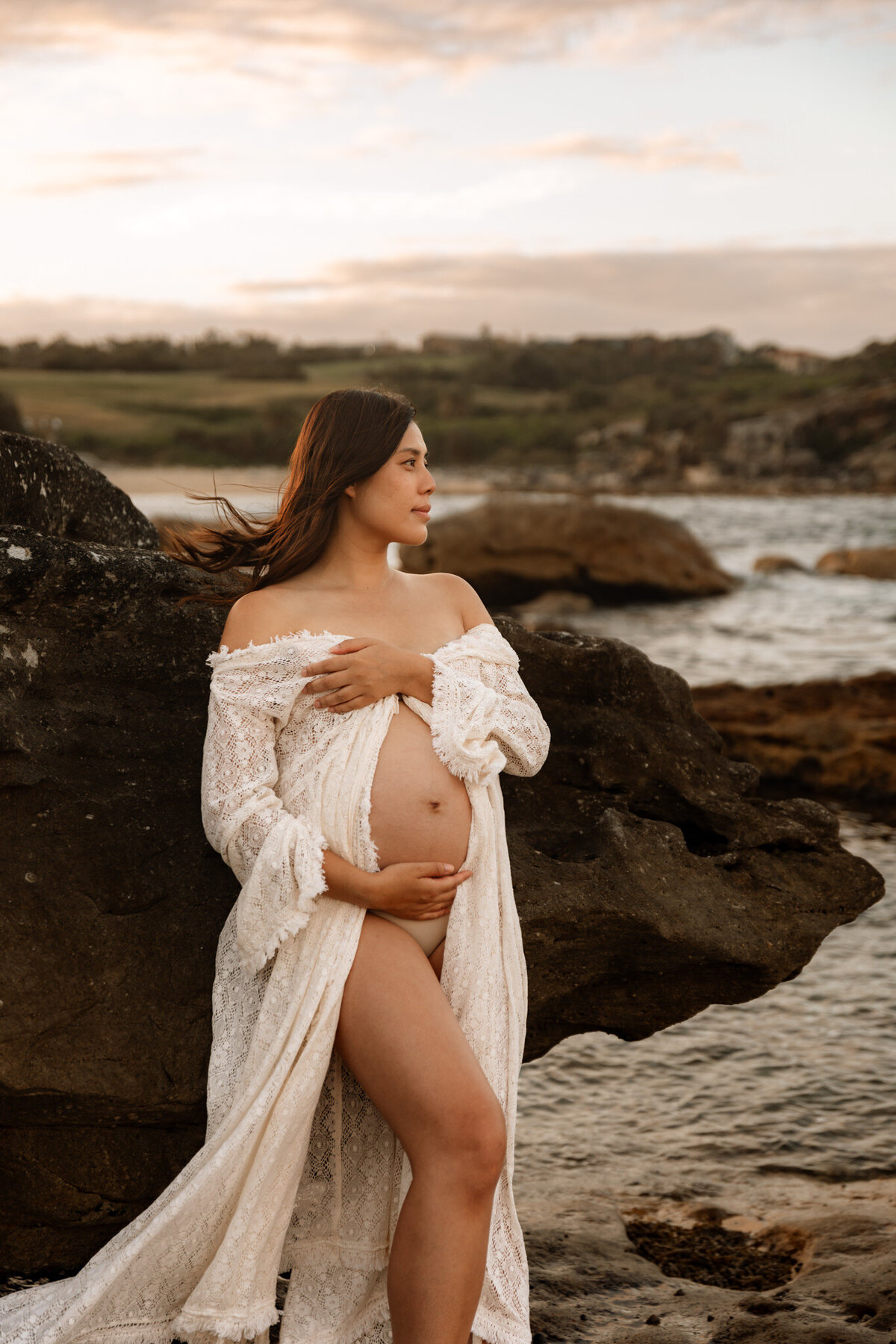 Expecting mother in lace robe cradling baby bump during a golden hour beach maternity session in Sydney with rocky coastal backdrop.