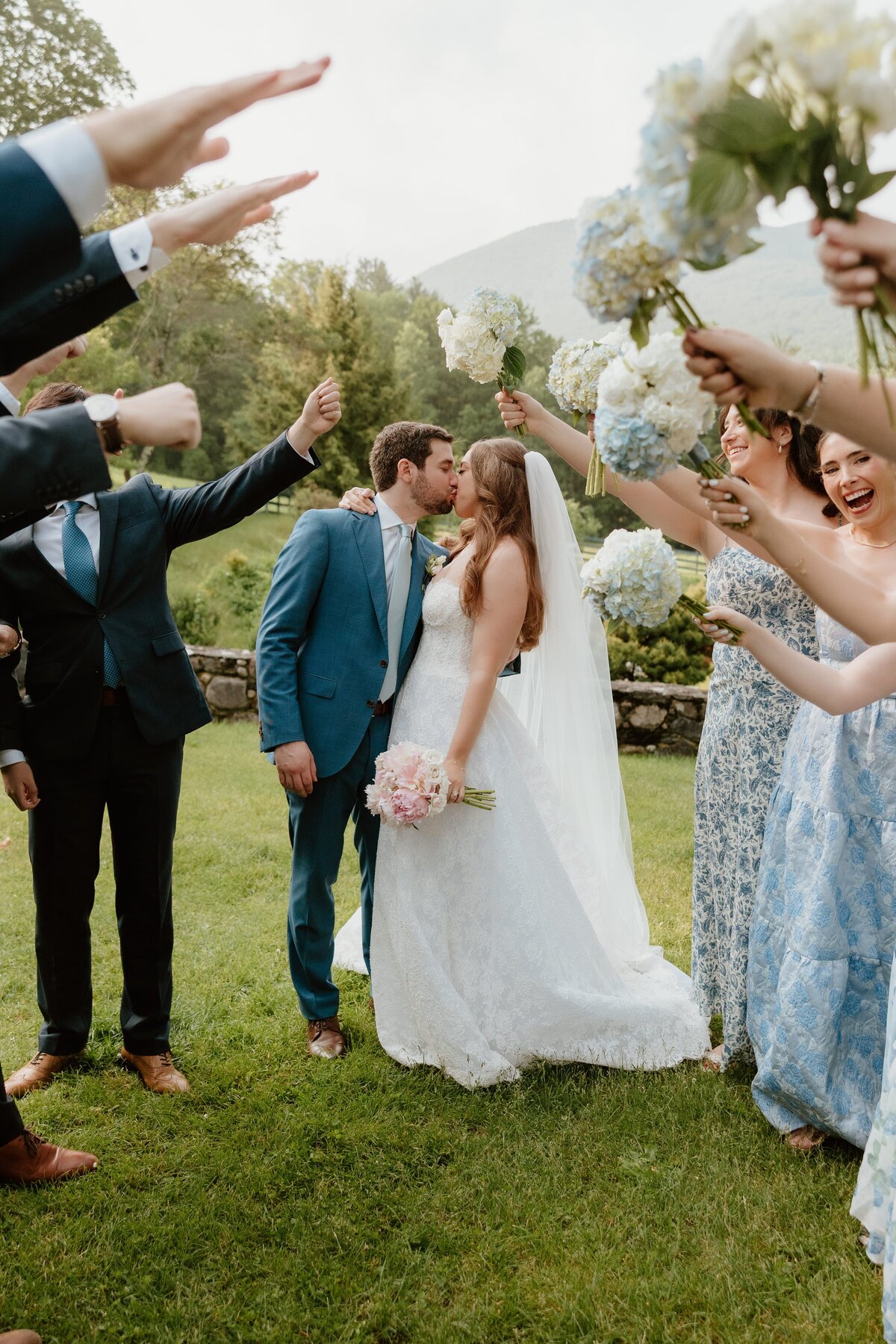 Newlyweds share a kiss as their wedding party celebrates around them with raised bouquets, creating a tunnel of love during a vibrant outdoor celebration.