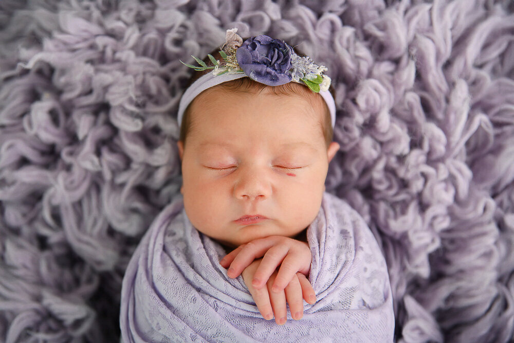 Newborn girl wrapped in purple laying on a purple rug.