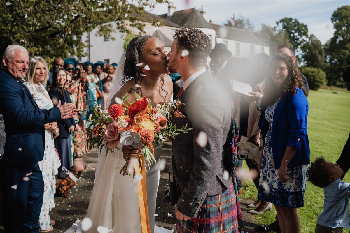 A bride and groom kiss between two lines of wedding guests as they throw confetti above them. Everyone smiles and laughs.. Logie Country House is photographed in the background and confetti blurs the lens.