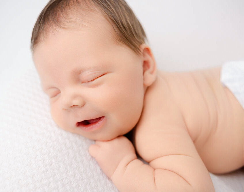 Newborn baby smiling and sleeping posed on belly with hand under chin.