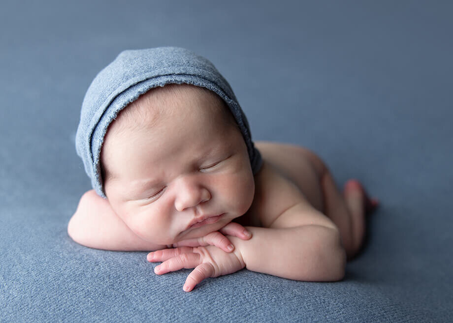 newborn boy posed chin on hands on top of denim blue fabric and he is wearing a matching beanie.
