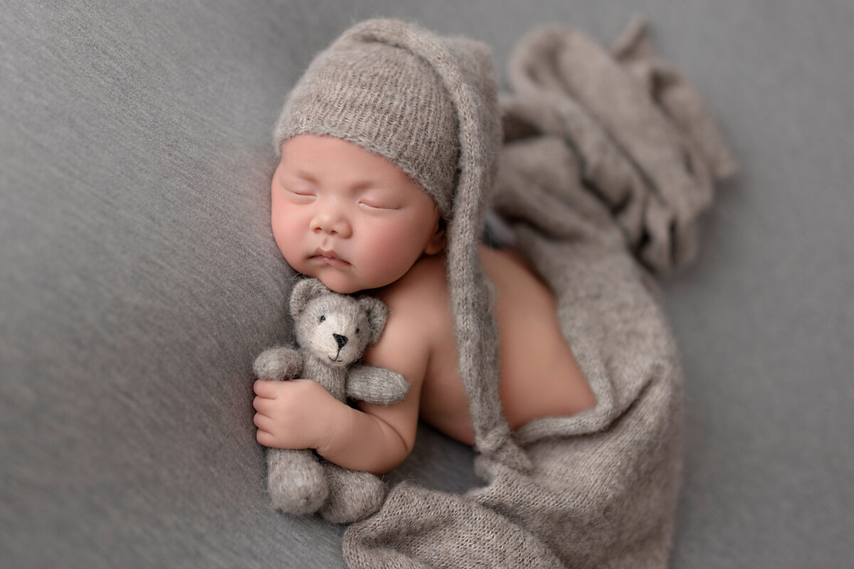 Newborn boy wrapped in grey knit fabric hugging a small teddy bear on a matching backdrop.