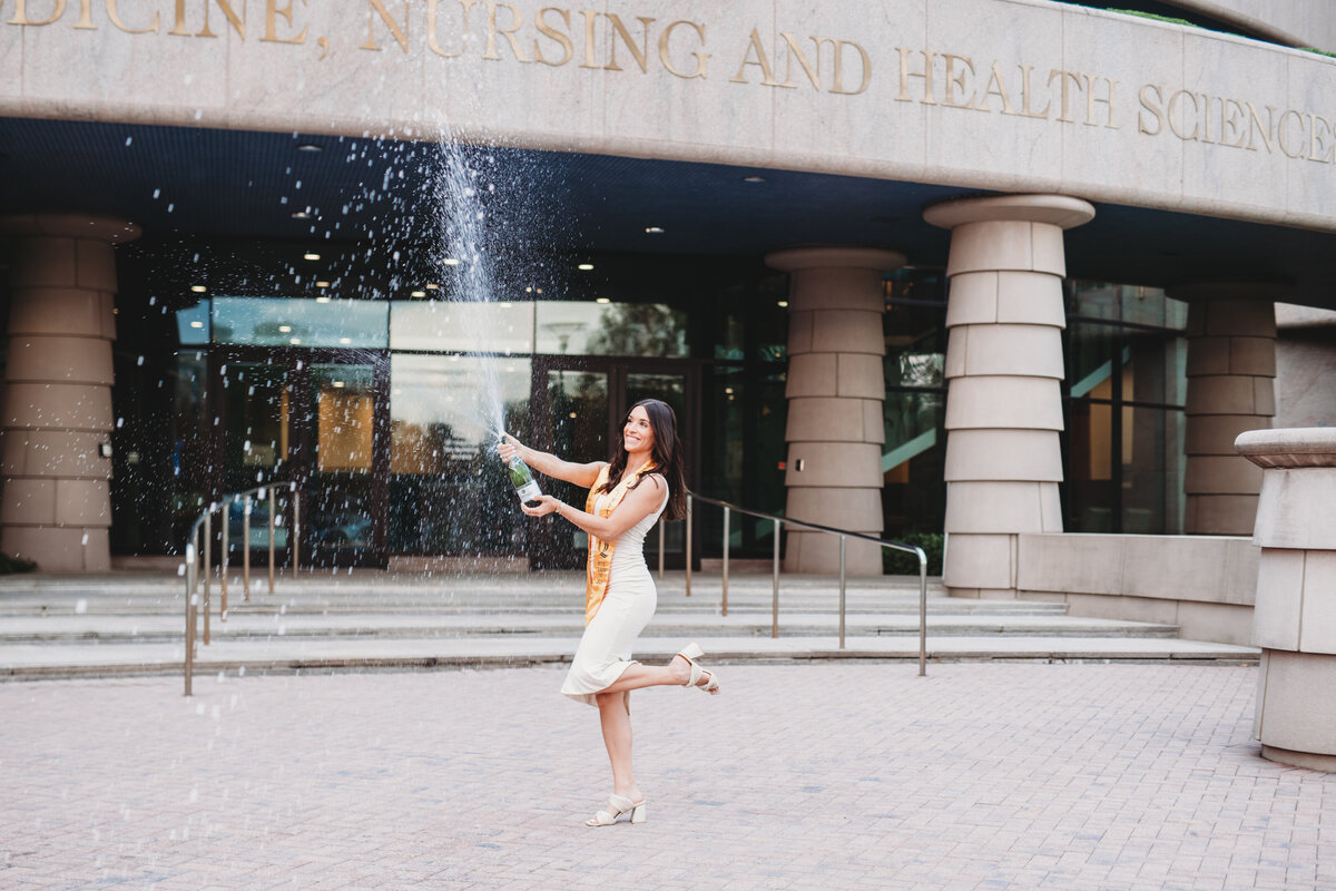 A joyful college graduate in a white dress and gold stole pops champagne in celebration outside a quinnipiac university building. Captured during a Connecticut graduation photography session by Nicole Mele Photography, highlighting achievement and excitement.