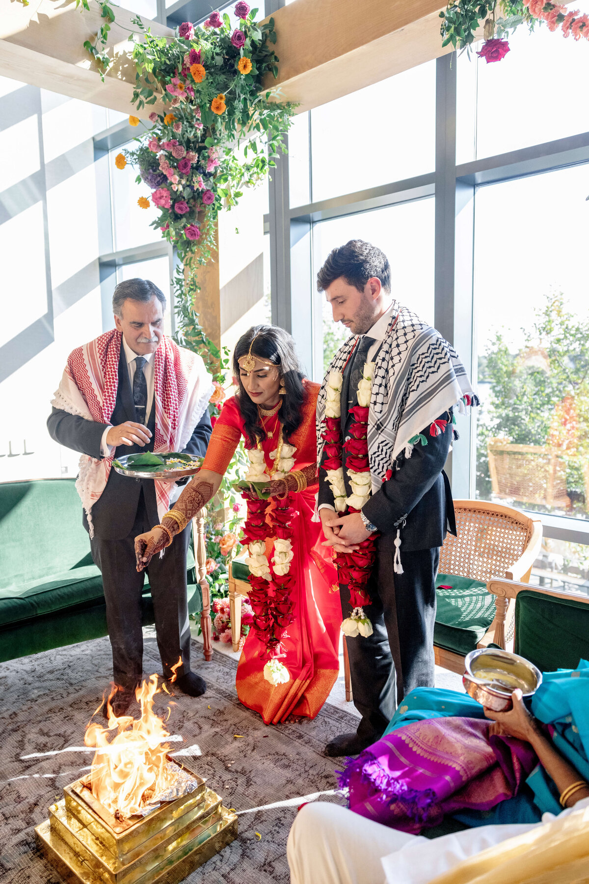 A couple stands at a traditional wedding ceremony, dressed in festive attire and floral garlands. They are participating in a ritual with fire, under a canopy adorned with flowers, with a priest assisting them in the ceremony.