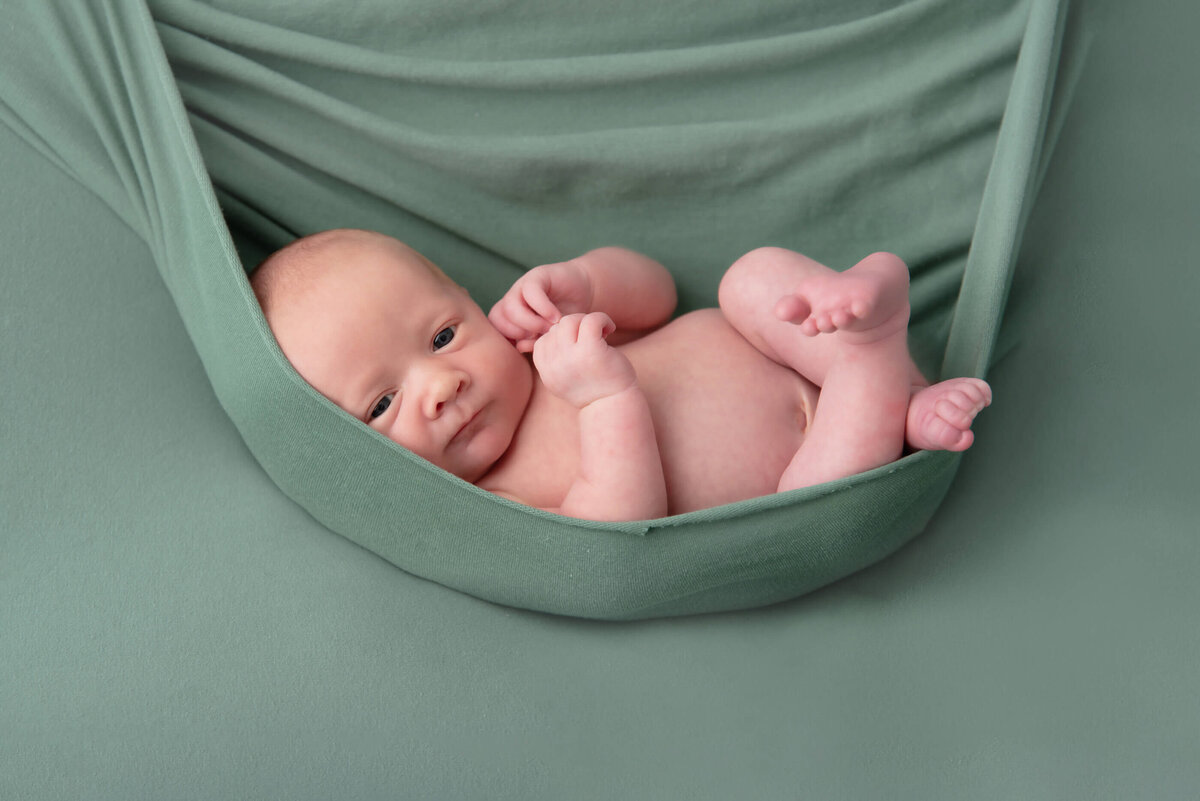 Newbonr baby boy with his eyes open in a green wrap on a green background for his newborn photos in Burlington, Ontario.