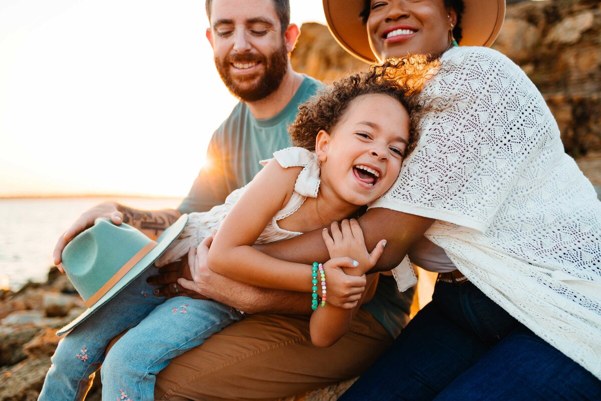 A joyful family enjoys a playful moment on the beach with their little girl. They are dressed in hats, blue jeans, and white shirts.