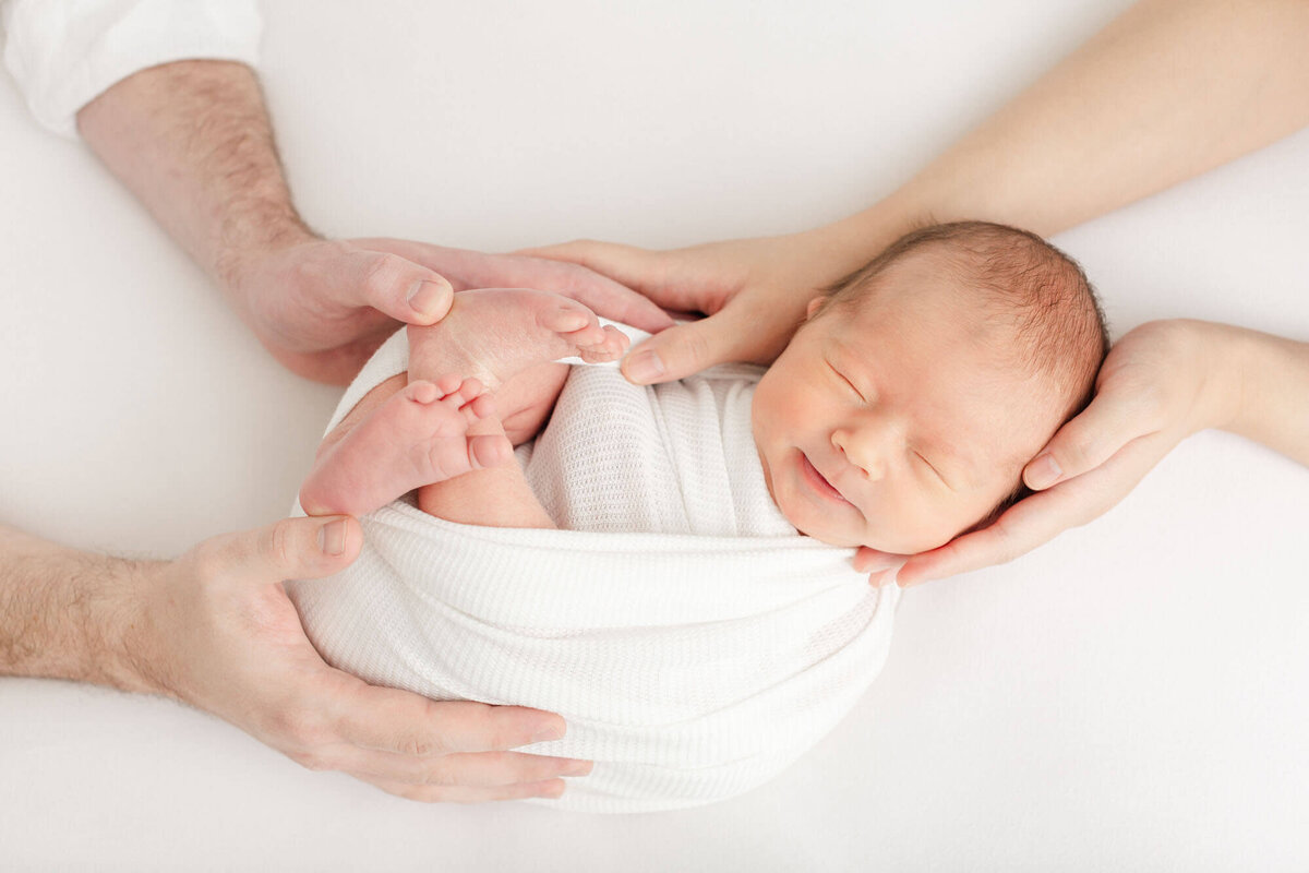 Baby is wrapped up in a white swaddle while mom and dad have their hands on him image taken from above.