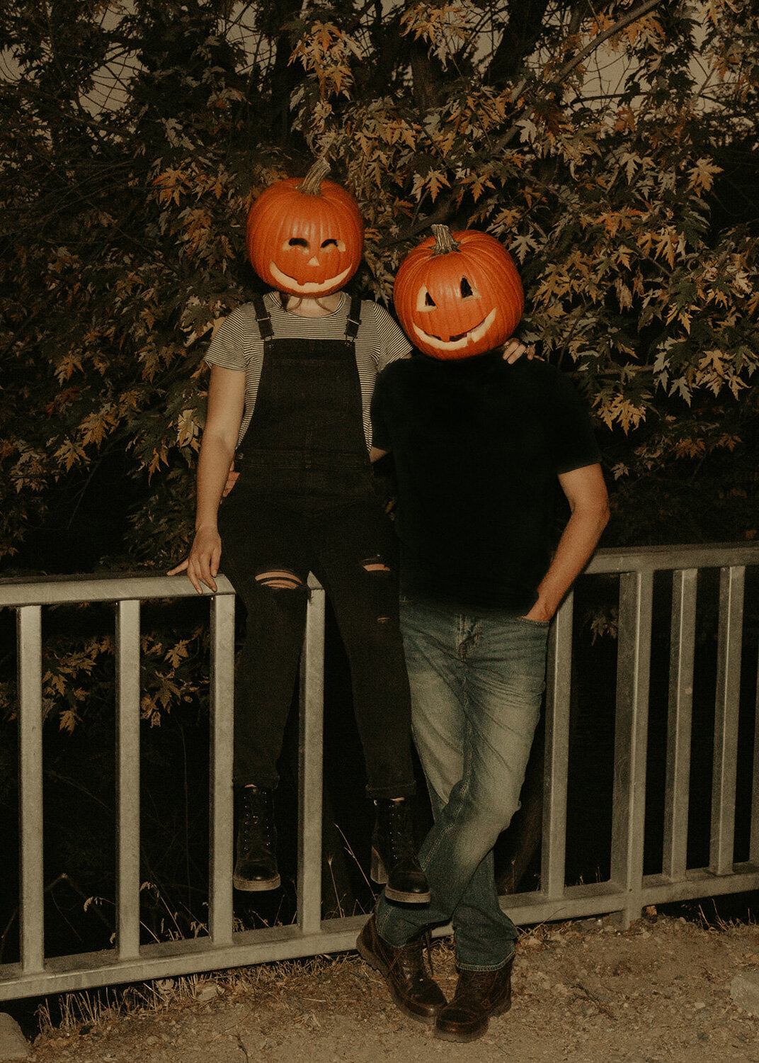 Couple wearing a pumpkin head while the man standing behind the railings and the woman sitting on the railings
