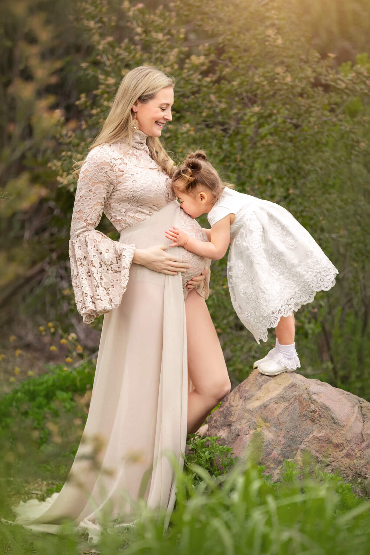 Pregnant mom wearing a beige dress with her toddler daughter standing on a rock and kissing moms baby bump