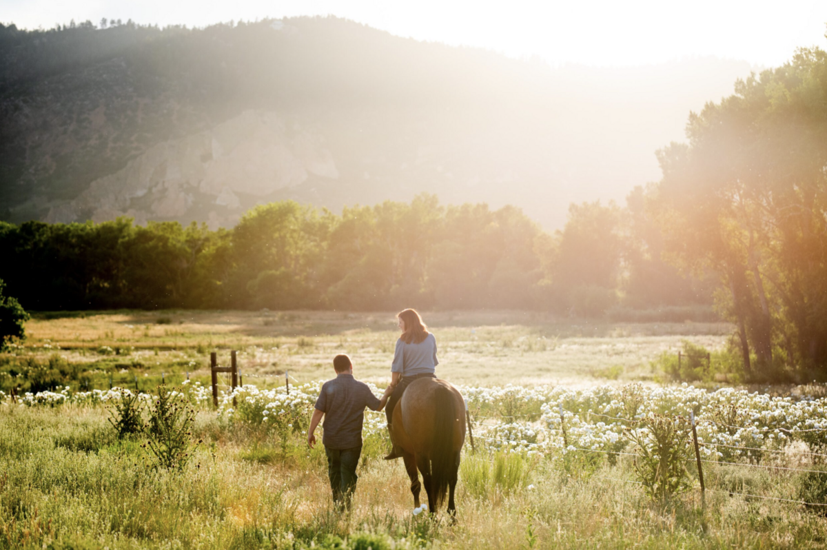 A leads a horse through a sunny field as his fiancée rides the horse, captured by Denver engagement photographer, Casey Van Horn.