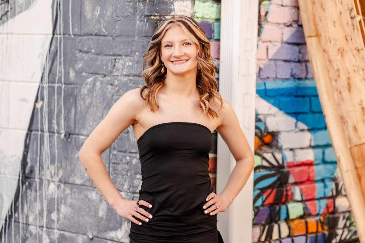 A high school senior in a black dress puts her hands on her hips and smiles. Behind her, you can see two walls with murals; one colorful and one monotone.